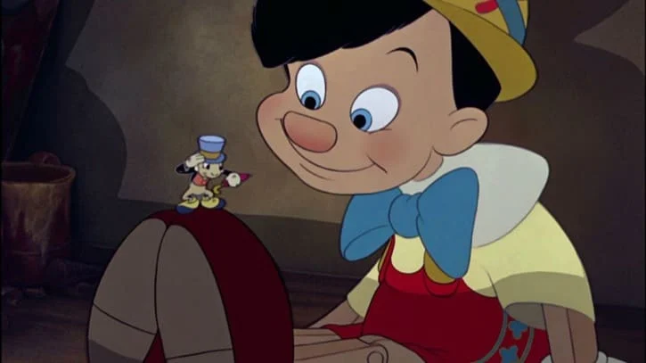 Pinocchio sitting down looking at Jiminy Cricket standing on the tips of his shoes